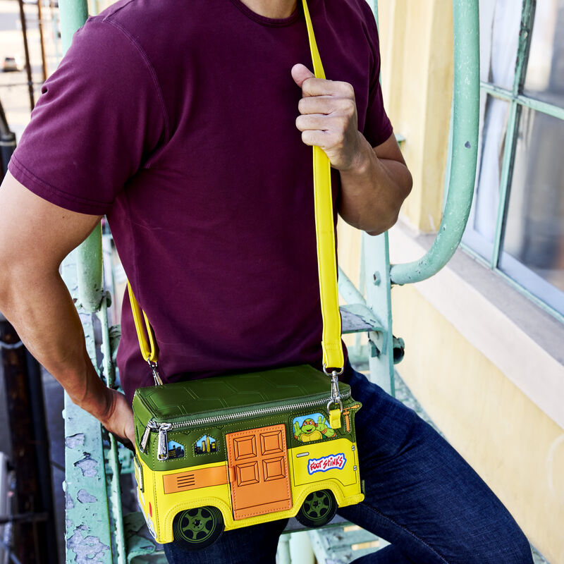 Man standing on a fire escape wearing a maroon shirt and blue jeans, wearing the Loungefly Teenage Mutant Ninja Turtles figural crossbody bag that looks like their party wagon.